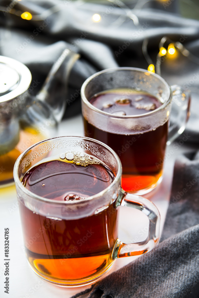 Black transparent teapot and cups with black tea on a wooden table next to a gray scarf and lights. Winter home concept Black transparent teapot and cups with black tea on a wooden table next to a gray scarf and lights. Winter home concept