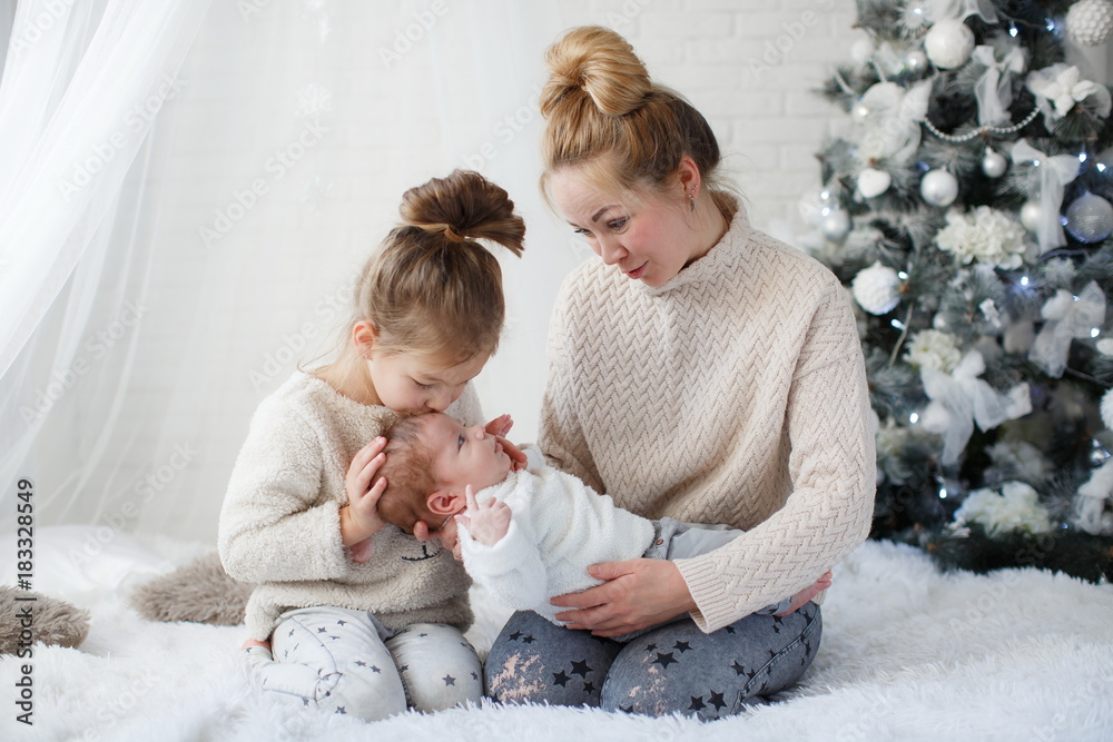 Happy woman,cheerful mother and little children,a girl and a newborn boy sitting on a fluffy white blanket beside the Christmas festive green Christmas tree with white toys,snowflakes,glowing lights Happy woman,cheerful mother and little children,a girl and a newborn boy sitting on a fluffy white blanket beside the Christmas festive green Christmas tree with white toys,snowflakes,glowing lights