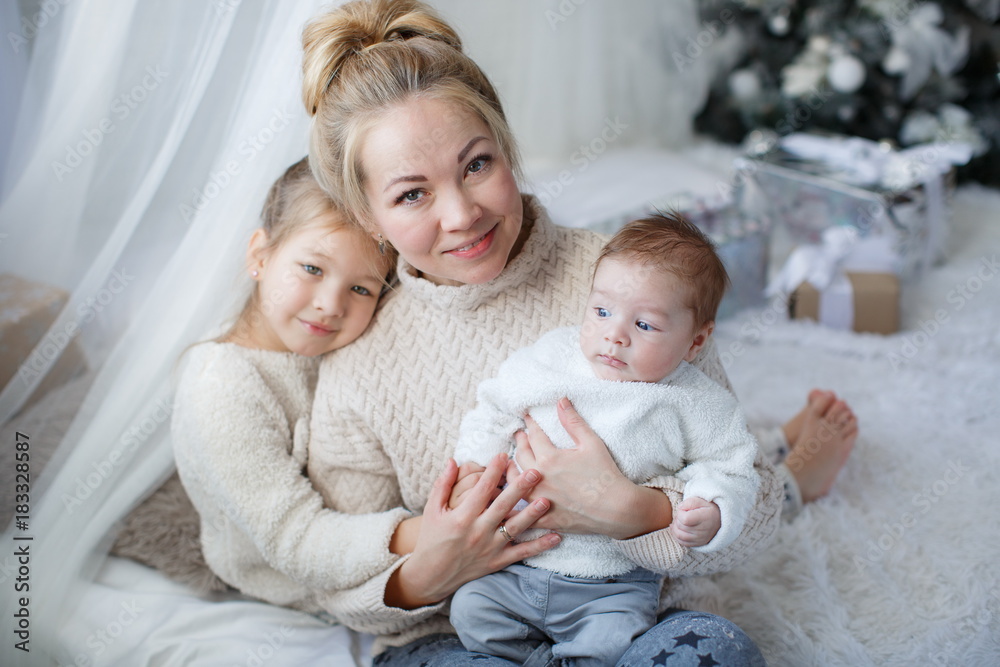 Happy woman,cheerful mother and little children,a girl and a newborn boy sitting on a fluffy white blanket beside the Christmas festive green Christmas tree with white toys,snowflakes,glowing lights