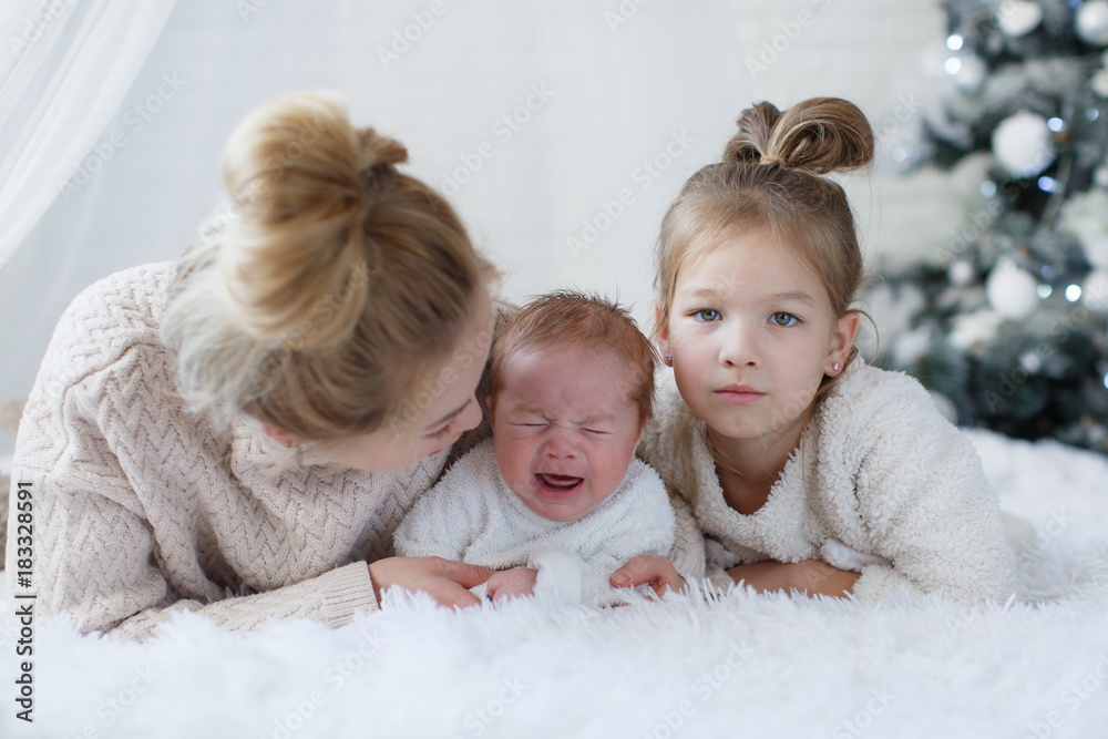 Happy woman,cheerful mother and little children,a girl and a newborn boy sitting on a fluffy white blanket beside the Christmas festive green Christmas tree with white toys,snowflakes,glowing lights Happy woman,cheerful mother and little children,a girl and a newborn boy sitting on a fluffy white blanket beside the Christmas festive green Christmas tree with white toys,snowflakes,glowing lights