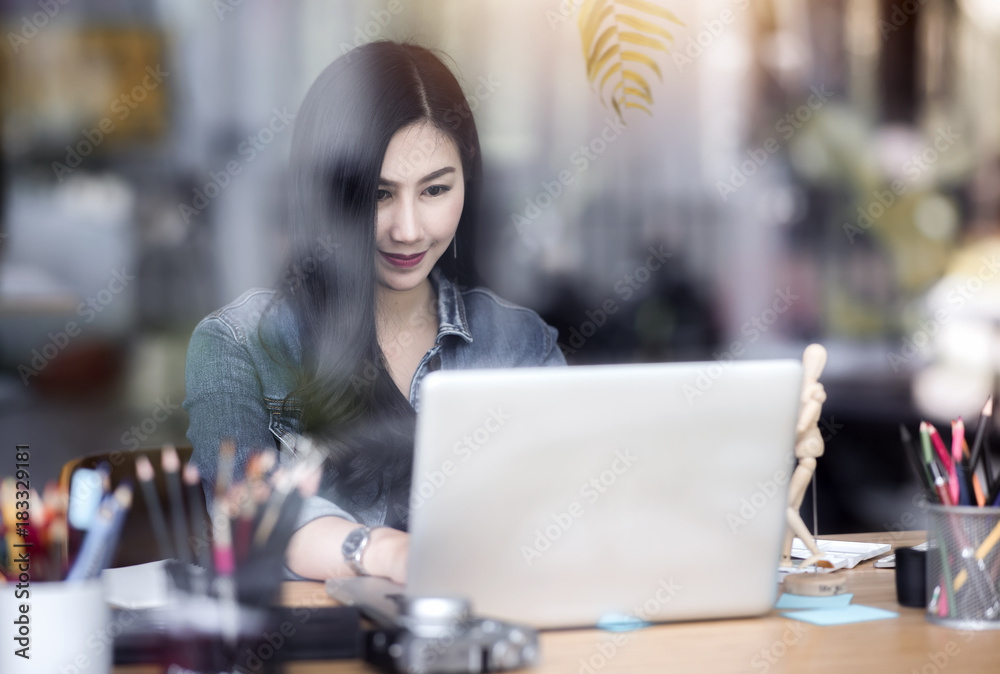 Smiling young beautiful woman designer sitting and using laptop in an office, looking through a window glass Smiling young beautiful woman designer sitting and using laptop in an office, looking through a window glass
