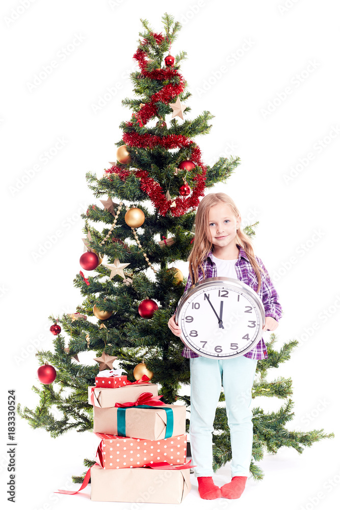 Portrait of little girl with clock showing five minutes to twelve standing near Christmas tree on white background Portrait of little girl with clock showing five minutes to twelve standing near Christmas tree on white background