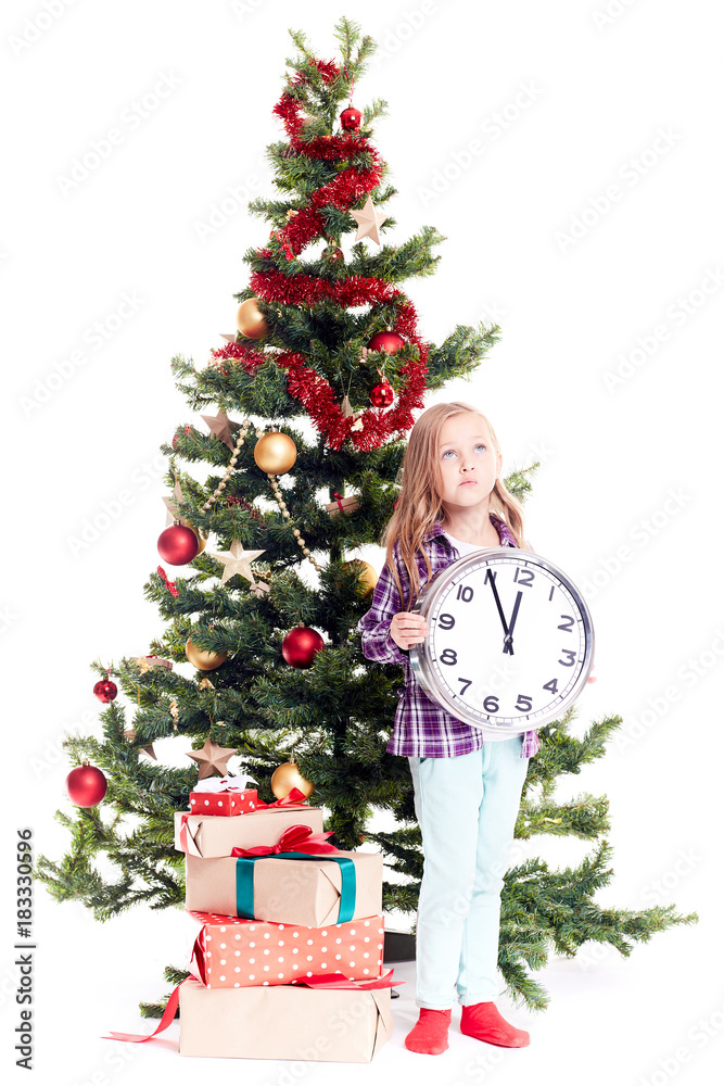 Portrait of little girl with clock showing five minutes to twelve standing near Christmas tree on white background Portrait of little girl with clock showing five minutes to twelve standing near Christmas tree on white background