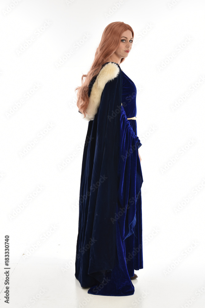 full length portrait of girl wearing long blue velvet gown and fur lined cloak, standing pose facing away from camera on white background.