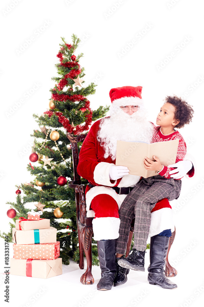 Santa Claus reading book for mixed-race little boy who is sitting on his lap near Christmas tree