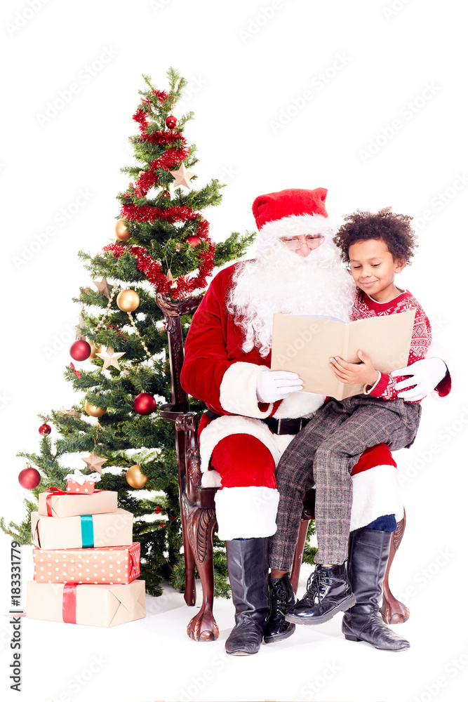 Santa Claus reading book for mixed-race little boy who is sitting on his lap near Christmas tree Santa Claus reading book for mixed-race little boy who is sitting on his lap near Christmas tree