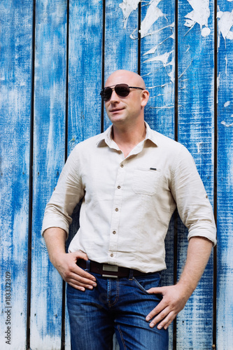 portrait of bald handsome man at natural shirt and jeans, boods. with sunglasses. model making pose at blue wood baghround. summer mood at spring time photo