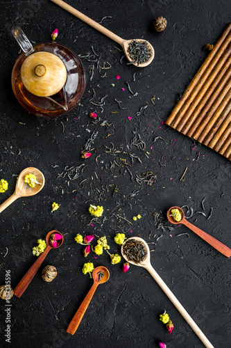 Brew the aromatic tea. Tea pot near wooden spoons with dried tea leaves, flowers and spices on black wooden background top view copyspace