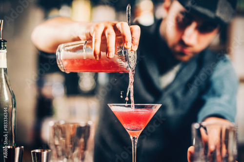 barman preparing and pouring red cocktail in martini class. cosmopolitan cocktail with bar background