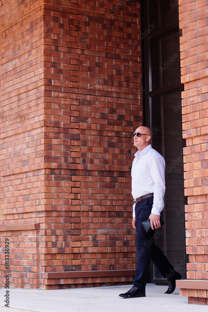 bald handsome businessman full height leaves the building red brick . shirt dark blue trousers, boots. spring, summer bald handsome businessman full height leaves the building red brick . shirt dark blue trousers, boots. spring, summer