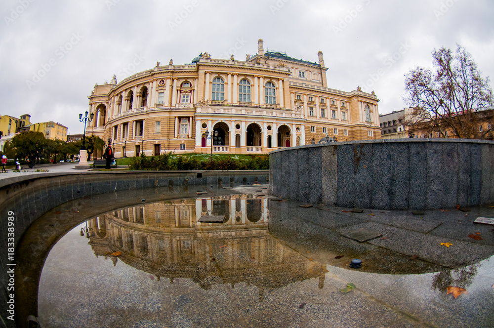 Square next to Odessa Opera House in autumn with grey empty stone fountain without water. Reflection of the building in the puddle