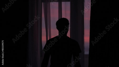 silhouette of a young man coming to a window and closing curtains against a window with a colorful sunset