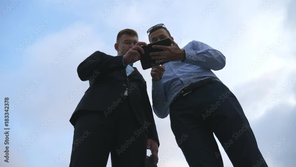 Two young businessmen talking and using tablet pc outdoor. Business men working on digital tablet outside with sky at background. Colleagues applying mobile technology. Low angle of view Slow motion Two young businessmen talking and using tablet pc outdoor. Business men working on digital tablet outside with sky at background. Colleagues applying mobile technology. Low angle of view Slow motion