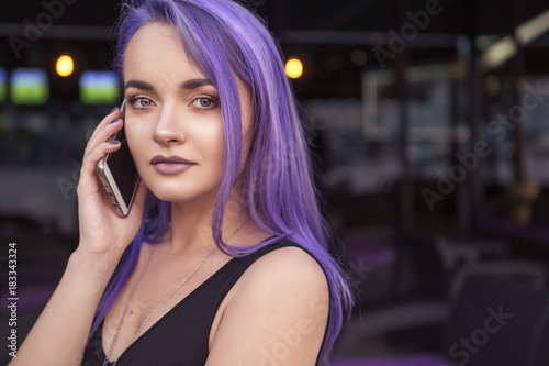 Beautiful brunette woman with colored (purple violet blue) hair in dark black slim dress outdoor in the european city, smiling, talking on a phone