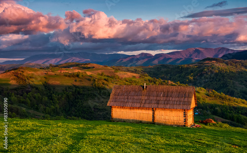 shed on the grassy hillside in red evening light. gorgeous springtime rural landscape in mountains under the blue sky with pink clouds