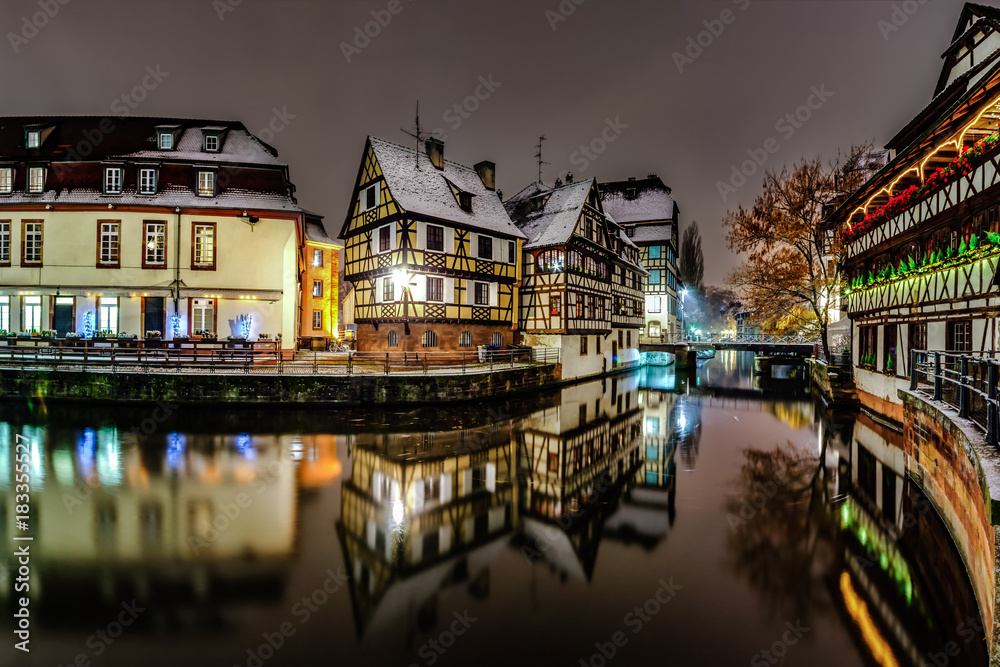 Old timber-framing houses in Petite France quarter, Strasbourg. Snow-covered roofs and refctions in the river water. Night scene.