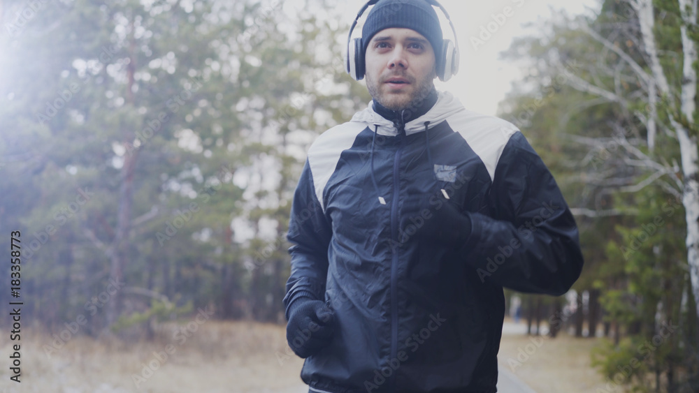 Fototapeta premium Attractive runner man in headphones jogging while listening music in winter park in the morning