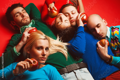 Picnic concept. Portrait of four stylish close friends hugging, having rest, holding red apples and lying on red background. Guys having fun. Hipster style. Studio shot © Augustino
