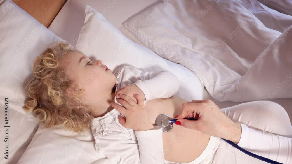 Pediatric doctor exams little cute girl lying in bed with stethoscope Pediatric doctor exams little cute girl lying in bed with stethoscope