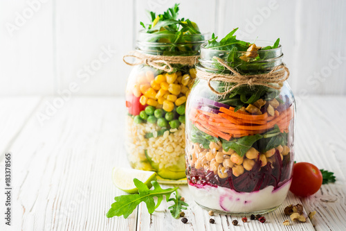 Healthy homemade salads with chickpeas, bulgur and vegetables in mason jars on white wooden background.