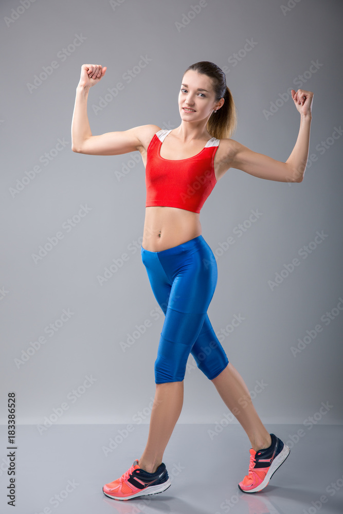 Smiling fitness girl flexing biceps and looking at camera. Three quarter length studio shot on gray background. Smiling fitness girl flexing biceps and looking at camera. Three quarter length studio shot on gray background.