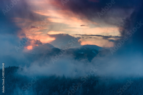 Landscape of dramatic sunset in the winter mountain. Wooded hills covered with snow  fog rising from valleys  colorful cloudy sky - this is impressive picture.  