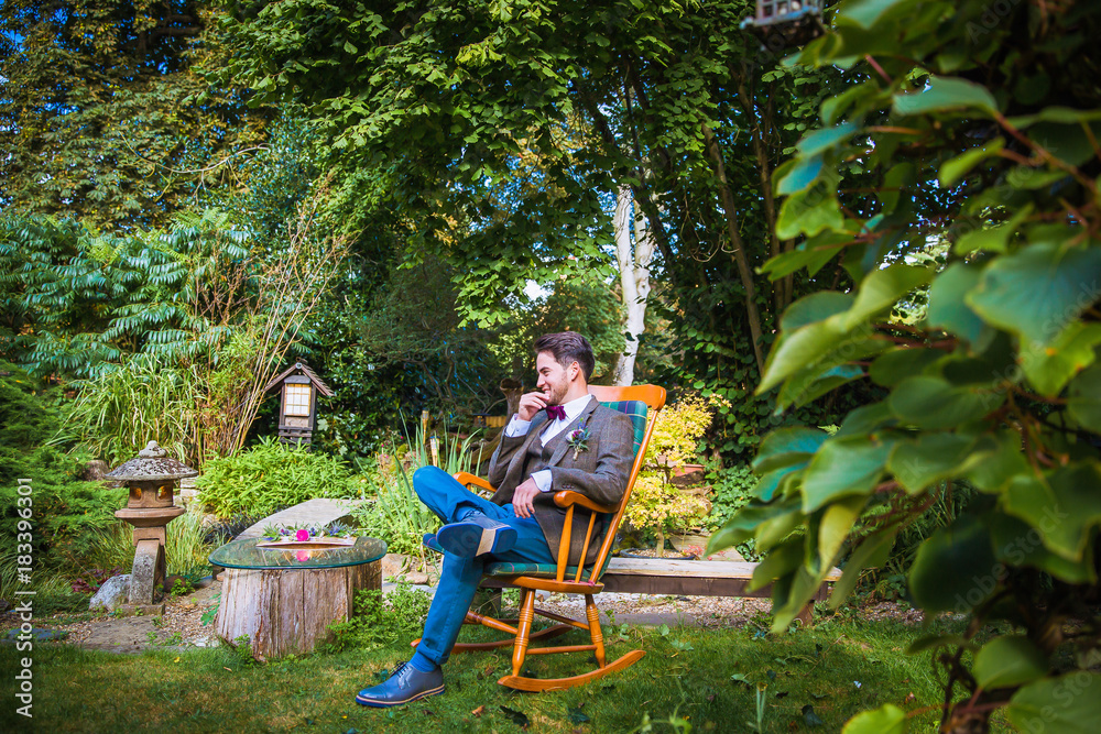 The young handsome smiling fiance sitting on the wooden chair in the garden