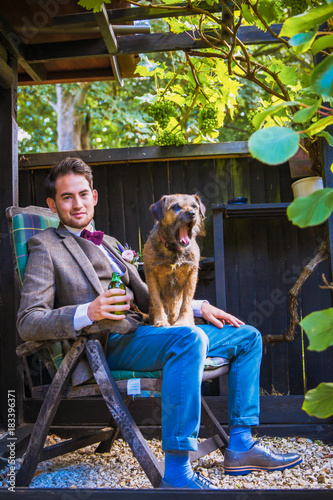 The young handsome joyful fiance sitting on the chair with his dog in the garden and holding bottle in hand