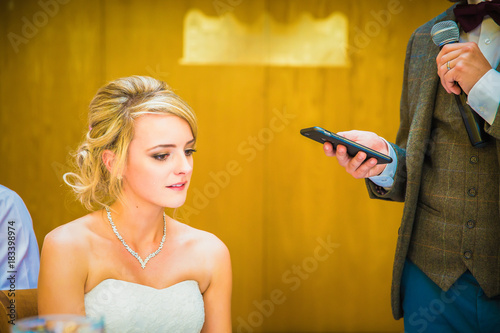 Portrait of the young beautiful bride who sitting by the table and her fiance standing near her and speaking in microphone in the restaurant