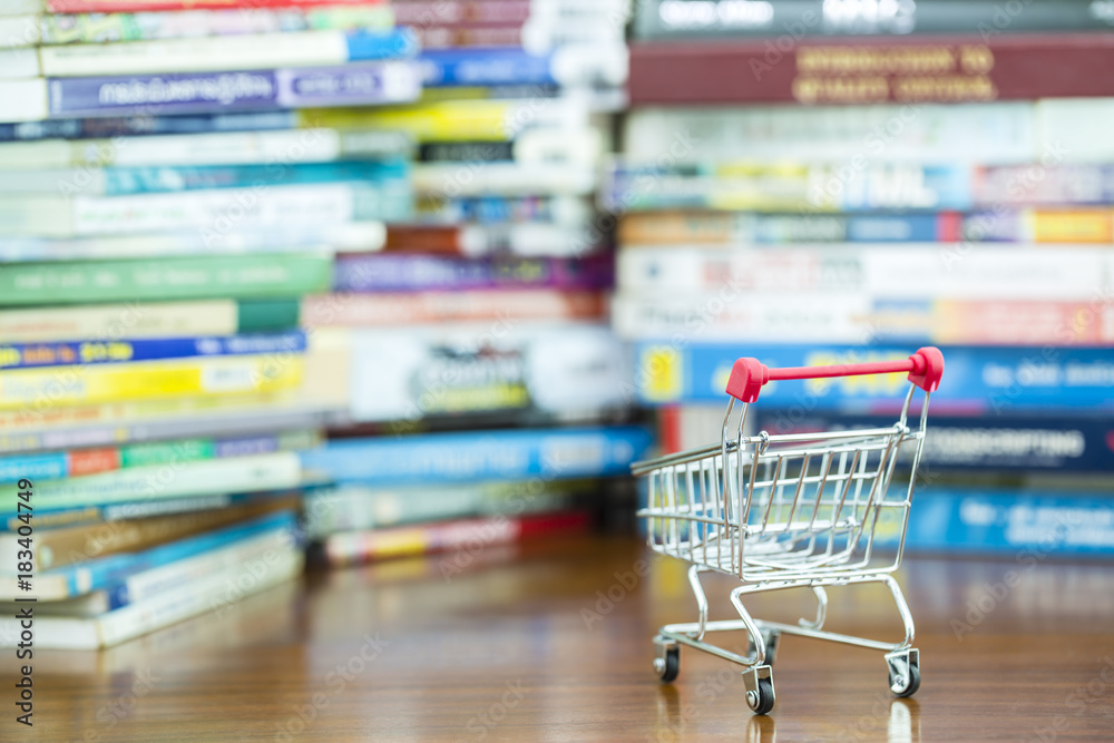 Mini shopping cart on wooden table with blurred pile of book as background, concept of buying knowledge to advance