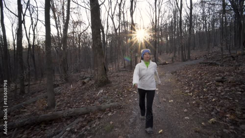 Mature blonde woman with a white jumper and blue headband hiking towards the camera down a mountain forest trail with the sun behind her. Mature blonde woman with a white jumper and blue headband hiking towards the camera down a mountain forest trail with the sun behind her.