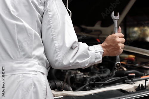 Professional young mechanic man in uniform holding wrench with open hood at the repair garage. Car insurance concept.