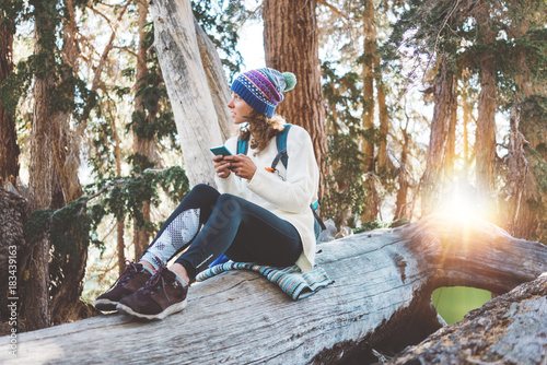 Traveling young woman with backpack, camp thermos and knitted hat typing on mobile phone text message and sitting on tree in wild forest at sunny day. Smiling.