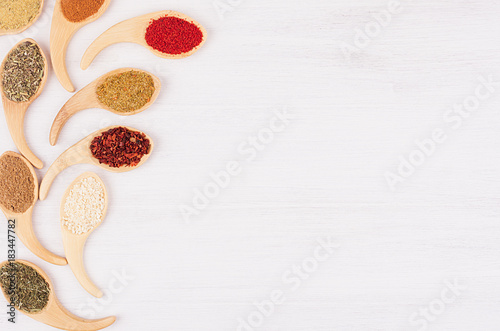 Decorative art border of different spices in bamboo spoons on white wood background, top view, closeup.