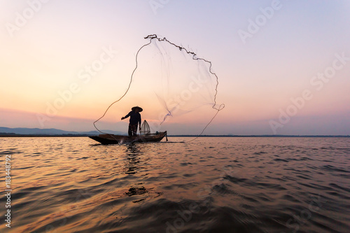 Thai fisherman on wooden boat casting a net for catching freshwater fish in nature river in the early evening before sunset.