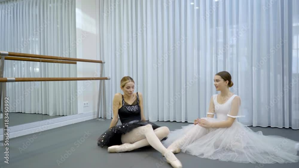 The two ballerinas sit on the floor in the bright studion during the break on repetition. The dancer in black tutu is leftside and the lady in white dress is on the right. The colleagues wears silk The two ballerinas sit on the floor in the bright studion during the break on repetition. The dancer in black tutu is leftside and the lady in white dress is on the right. The colleagues wears silk