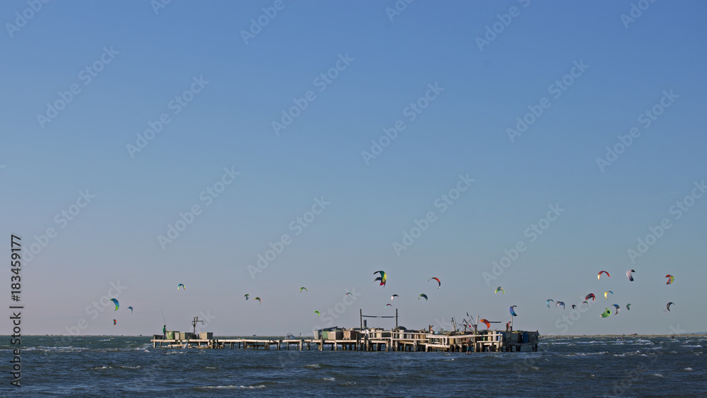 Kitesurf, parapente et voile delta au-dessus de la mer Méditerranée sur une plage de Camargue avec ponton de pêche.