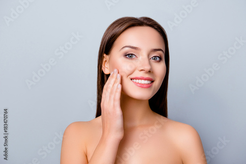 Close up photo of lovely attractive smiling woman with shiny smile, she is touching her cheek after using cream, isolated on grey background