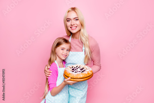 Studio portrait of happy mother and daughter holding together backed pie with jam inside for father, looking to camera, standing over pink background