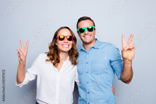 Cute, cool, lovely, funny, positive partners in colorful summer glasses, shirts, casual outfit smiling to the camera and showing peace symbol standing  over grey background