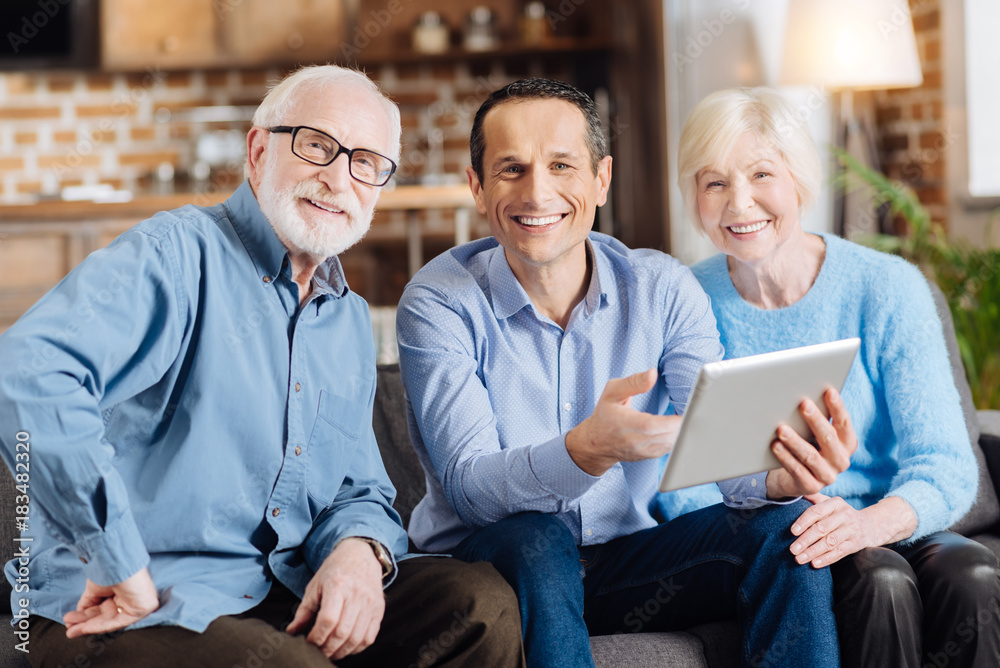 Happy students. Cheerful young man sitting on the sofa between his elderly parents and smiling at the camera together with them while teaching them how to use a tablet Happy students. Cheerful young man sitting on the sofa between his elderly parents and smiling at the camera together with them while teaching them how to use a tablet