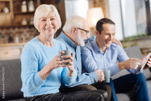 Feeling cozy. Charming elderly woman posing with a coffee cup and smiling while her husband and son using a tablet together in the background