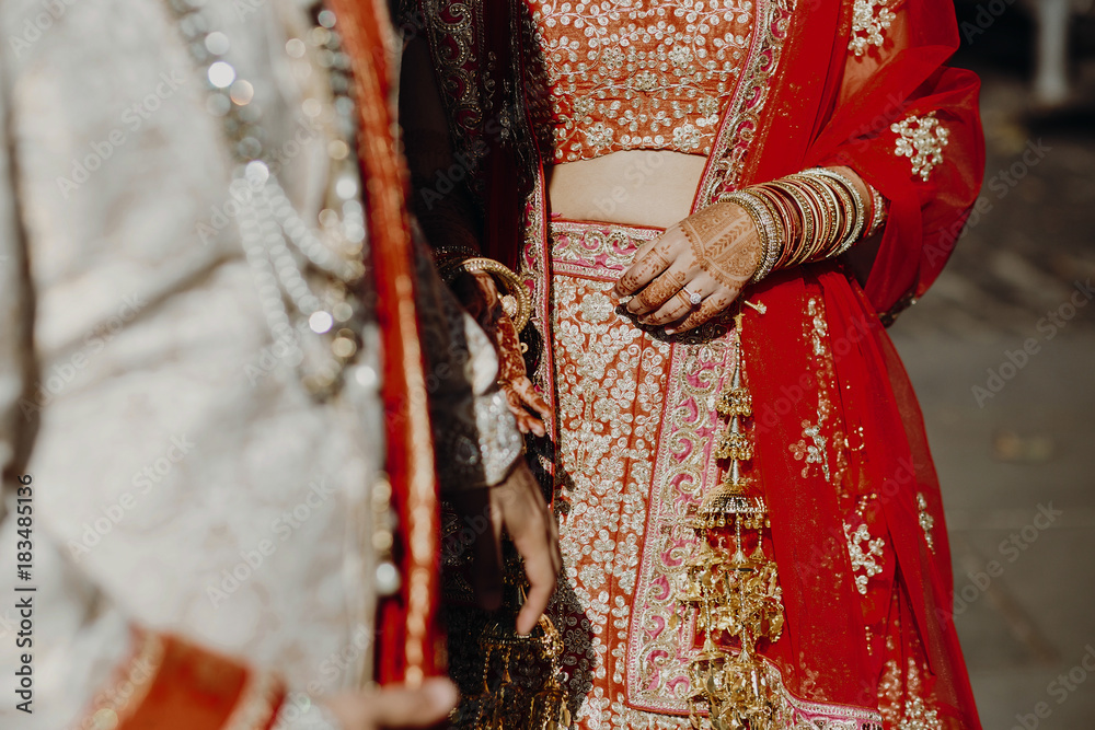 Indian groom dressed in white Sherwani and red hat with stunning bride in red lehenga stand and hold each hands walking outside