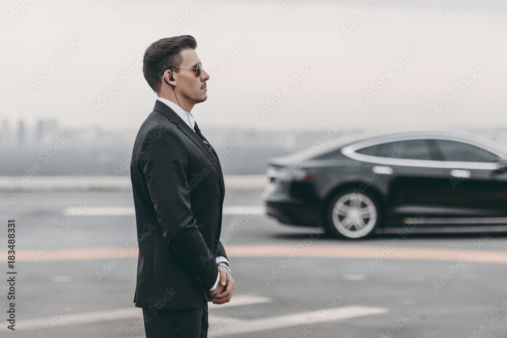 bodyguard in suit with security earpiece standing close to politician car