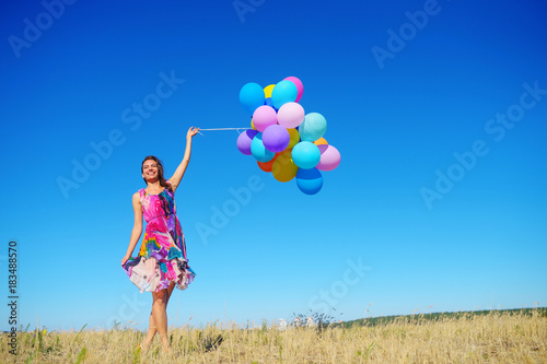 Beautiful, cheerful young woman in a bright dress with colorful balloons on a field with a blue sky in the summer. Warm sunny summer day with light breeze.