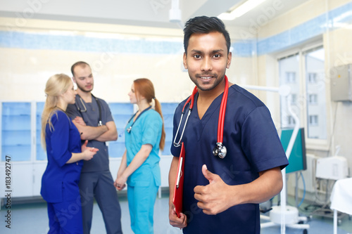 Asian male doctor in front of team, looking at camera, show thumb up, with medical team in background. Multiracial team of young doctors in a hospital standing in a operating room.