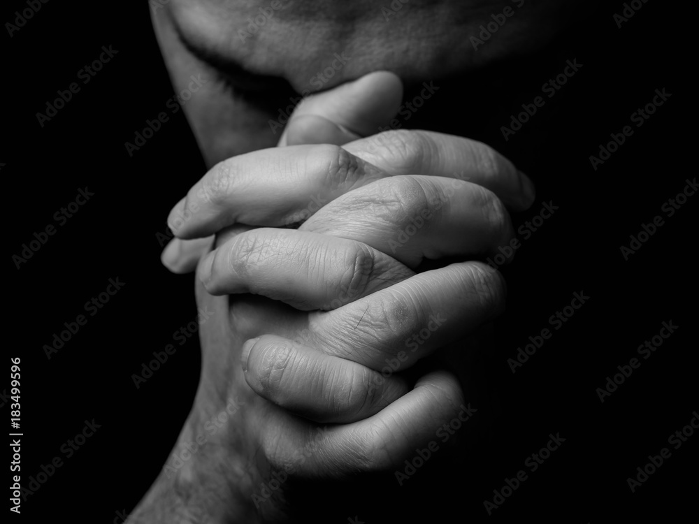 Close up of faithful mature man praying, hands folded in worship to god with head down and eyes closed in religious fervor. Black background. Concept for religion, faith, prayer and spirituality. - obrazy, fototapety, plakaty 