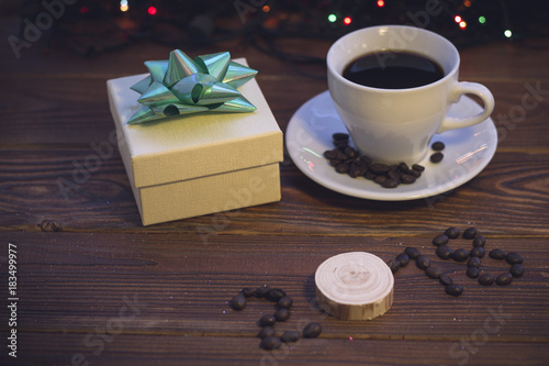 A romantic festive still life with a cup of coffee with, coffee beans on a saucer, a beige gift box with a green bow and 2018 inscription of coffee beans on a rustic wooden background.