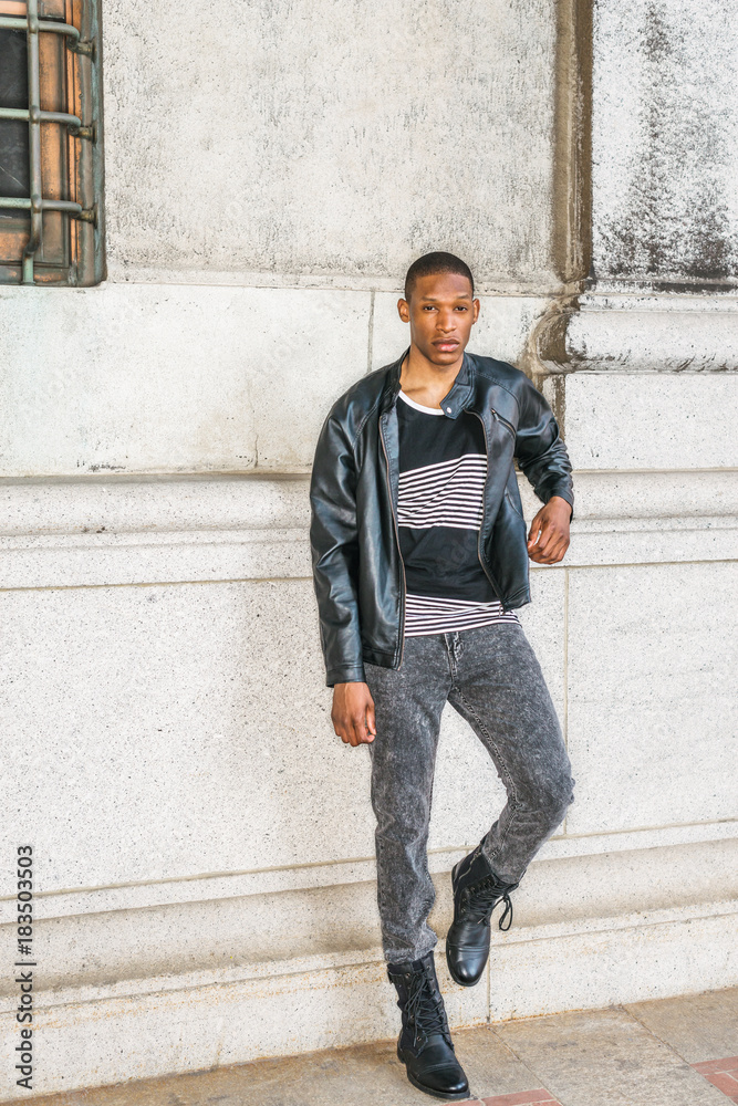 Man Urban Casual Fashion. Wearing black leather jacket, striped undershirt,  jeans, leather boots, a young African American guy standing against wall on  street in New York, relaxing, waiting for you.. Stock Photo