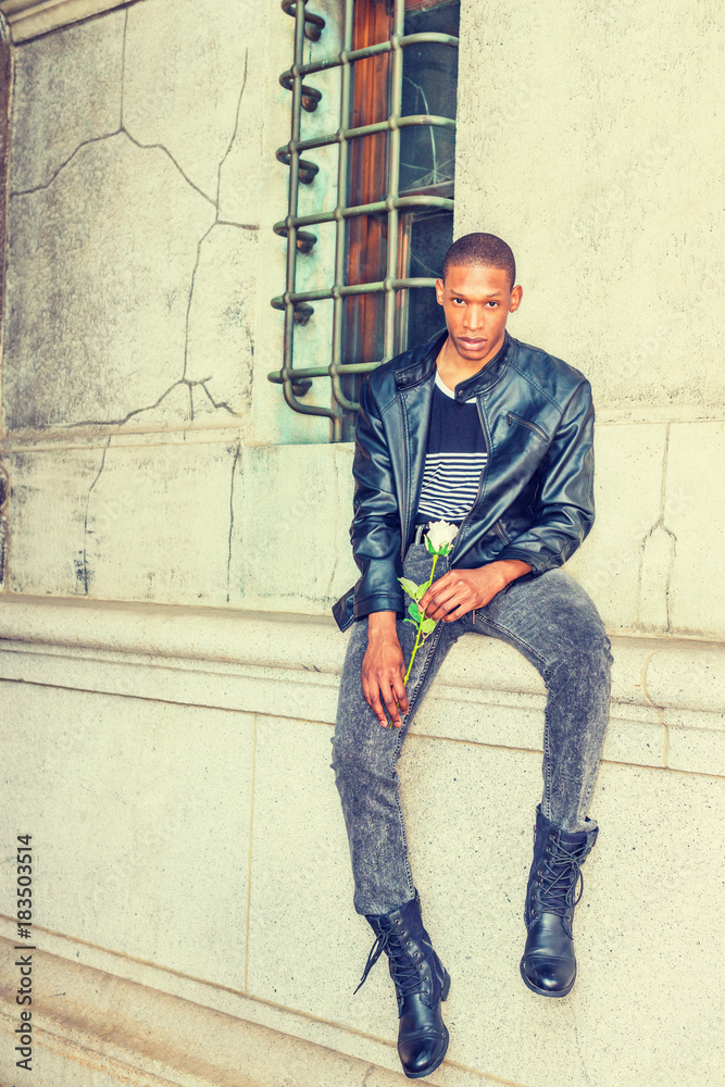 Stockfoto Man seeking love in New York. Wearing black leather jacket,  striped undershirt, jeans, boots, hands holding white rose, a young black  guy sitting against wall with window on street, waiting for